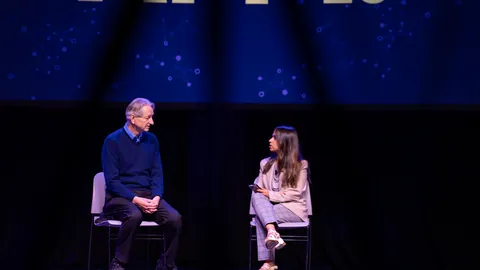 A man and a woman talking on a blue-lit conference stage.