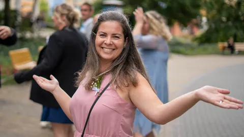 A smiling wedding guest in a pink dress with her arms outstretched.