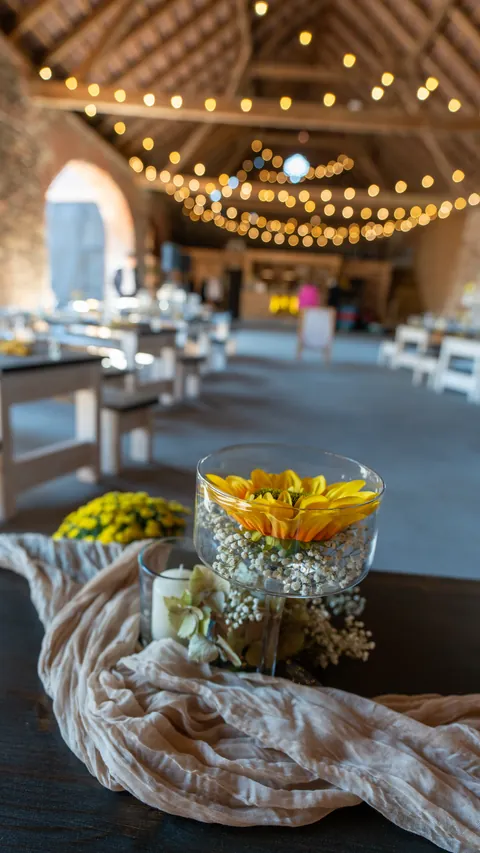 A yellow flower in a glass vase and string lights in a wedding barn.